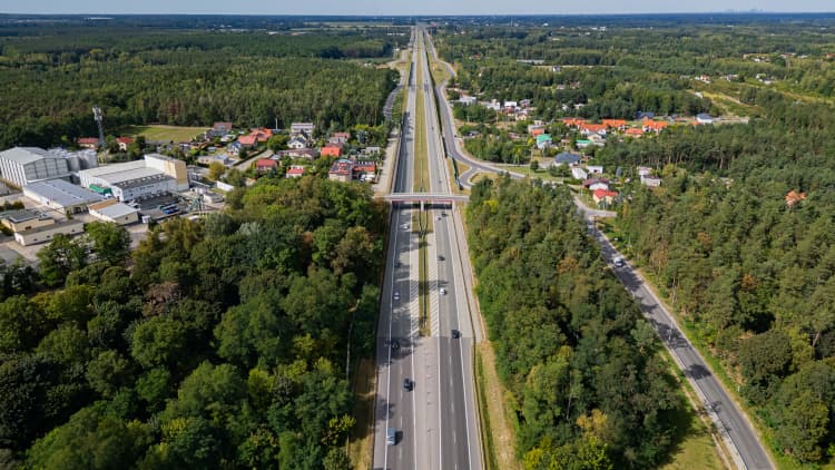 Autostrada przecinająca zalesiony teren z mostkiem nad jezdnią i zabudową przy lesie